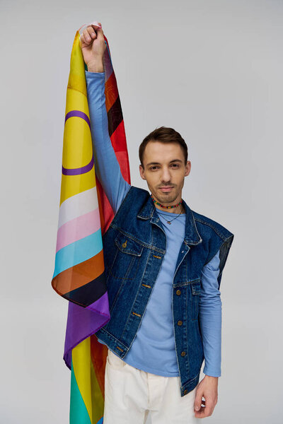 merry handsome gay man in vibrant casual attire holding rainbow flag and smiling at camera