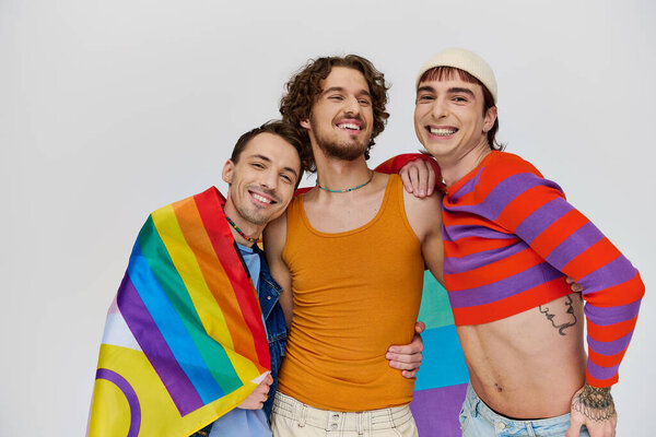three joyous stylish gay men in cozy clothing posing actively with rainbow flag on gray backdrop