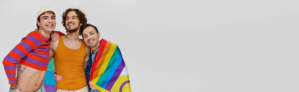 three jolly gay men in cozy clothing posing actively with rainbow flag on gray backdrop, banner