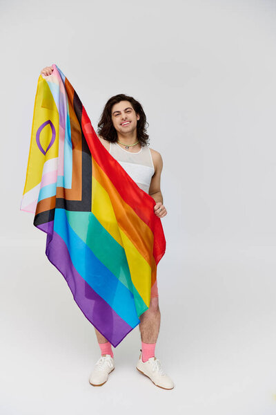 handsome joyous gay man with long dark hair posing with rainbow flag and looking at camera
