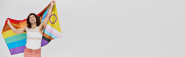 joyous fancy gay man with long dark hair posing with rainbow flag and looking at camera, banner