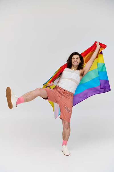alluring joyous gay man with long dark hair posing with rainbow flag and looking at camera