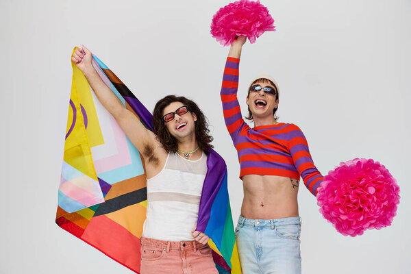 two joyful alluring gay men in bold attires with sunglasses posing with rainbow flag and pom poms