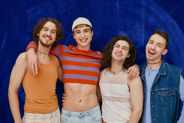 four positive appealing stylish gay friends in everyday bright attires on blue backdrop, pride month