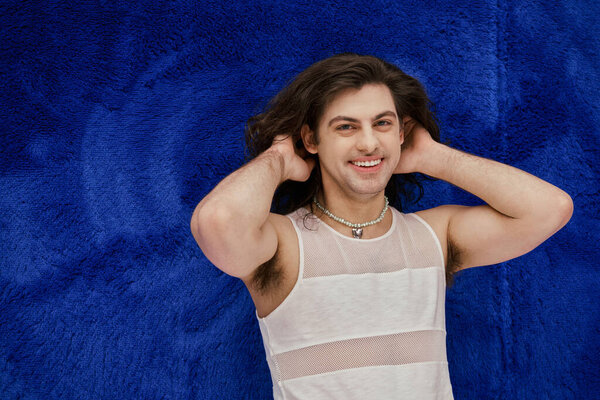 handsome cheerful gay man with long hair smiling at camera on dark blue backdrop, pride month