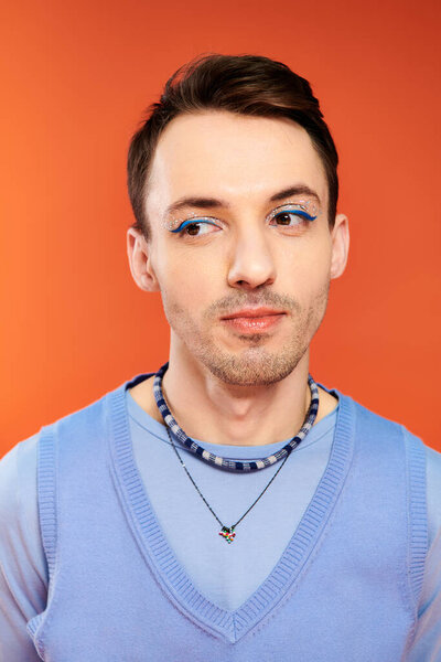 contented attractive stylish gay man with vibrant makeup posing on orange backdrop, pride month