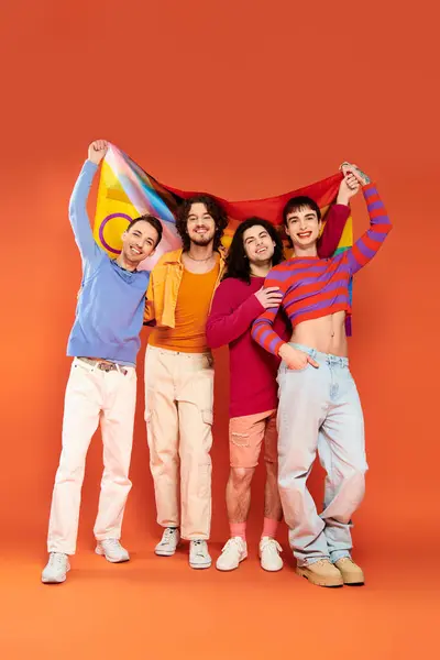 カメラの前に虹の旗を掲げている活気に満ちた服装の4人の楽しいファッショナブルなゲイ男性 — ストック写真