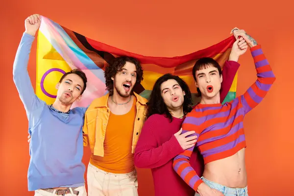 カメラの前に虹の旗を掲げている活気に満ちた服装の4人の楽しいファッショナブルなゲイ男性 — ストック写真