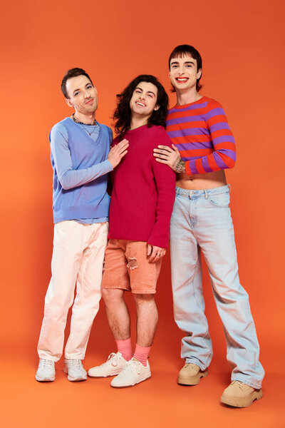 three well dressed joyous gay friends in vivid clothes posing together on orange backdrop, pride