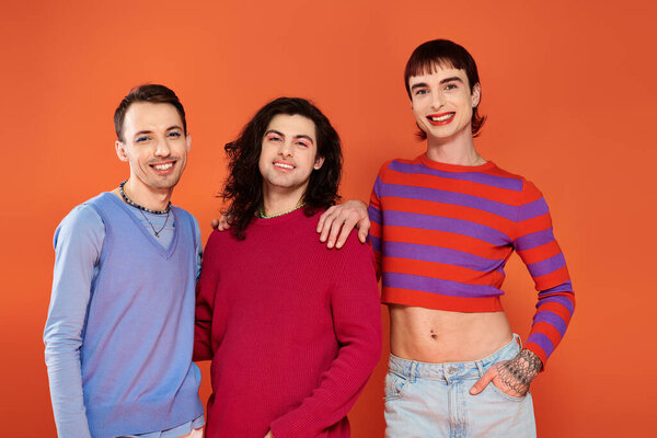 three fashionable joyous gay friends in vivid clothes posing together on orange backdrop, pride