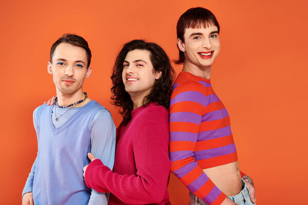 three sophisticated joyous gay friends in vivid clothes posing together on orange backdrop, pride