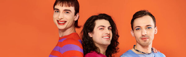 three jolly gay friends in vivid clothes posing together on orange backdrop, pride month, banner