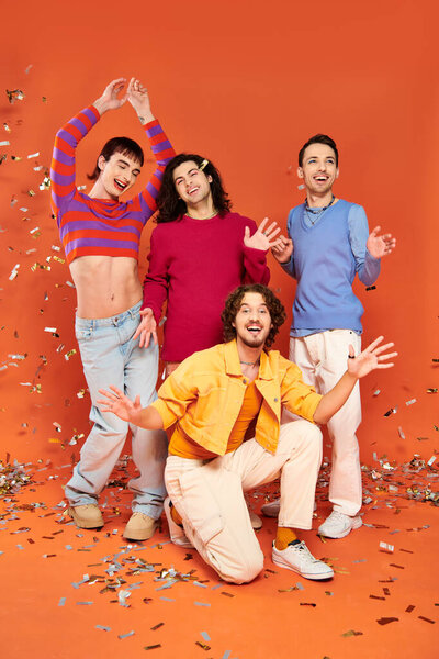 four cheerful stylish gay friends in bold outfits posing actively under confetti rain, pride month