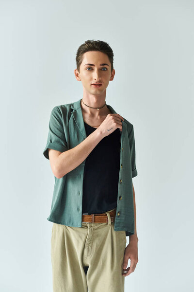 A young queer person confidently posing in a studio, wearing a stylish green shirt and tan pants on a grey background.