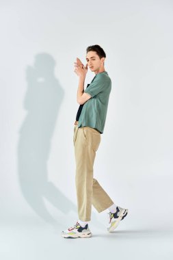Yeşil tişörtlü ve haki pantolonlu genç bir eşcinsel stüdyoda beyaz bir duvarın önünde güvenle duruyor..