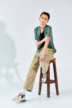 Genç ve tuhaf bir insan, gri bir arka plana karşı gururlu ve güçlü bir şekilde stüdyoda bir tabureye oturur..