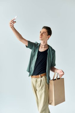 Genç bir eşcinsel alışveriş çantası tutuyor, stüdyoda gri bir arka planda selfie çekiyor..