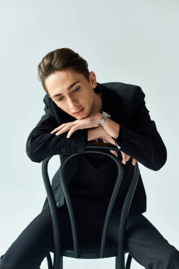 Genç bir ibne, siyah bir takım elbiseyle gri bir stüdyonun arka planında otururken zarafet yayar..