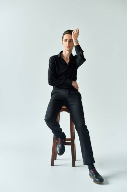 Genç, eşcinsel bir adam beyaz bir arkaplanın önündeki taburede huzur içinde oturur. Zarafet ve kendine güven yayar..