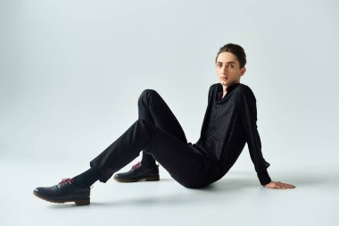 A young queer person is seated on the floor, wearing a black sweater, striking a pose in a studio with a grey background. clipart