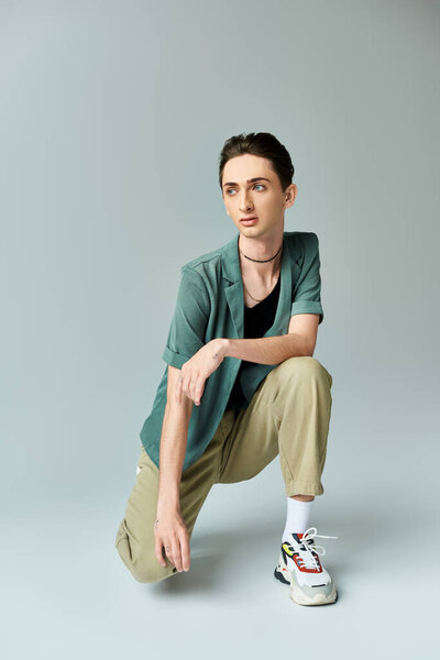 A young queer person crouching down in a pose of contemplation against a grey background in a studio setting.