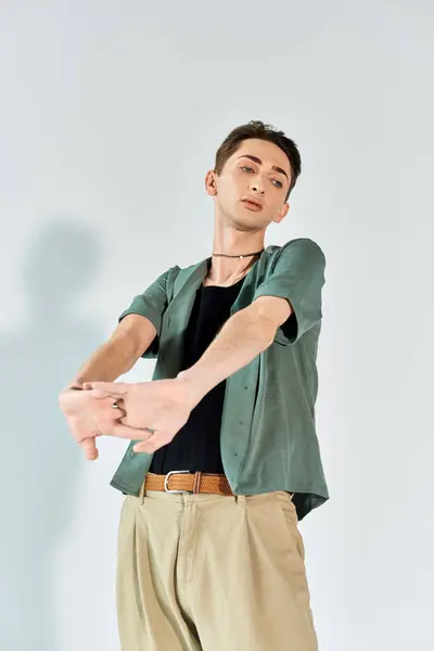 stock image A young queer person striking a confident pose in a studio, wearing a green shirt and tan pants against a grey background.