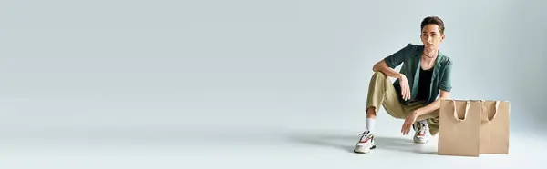 stock image A young queer person crouching down with shopping bags in a studio on a grey background.