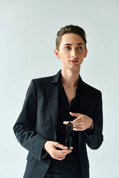 A young queer person in a stylish black suit strikes a confident pose against a grey studio backdrop.