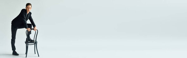 A young queer person leans on a chair in a studio against a white background, striking a contemplative pose.