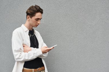Stylish young queer person in casual attire leaning against a wall, holding a cell phone. clipart