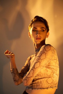 A young queer person in a snakeskin top strikes a pose in front of a bright light, exuding style and confidence. clipart