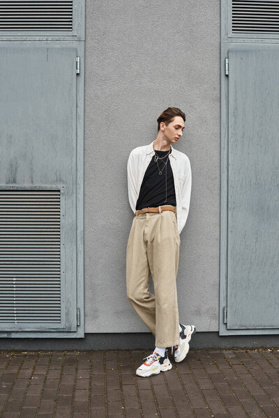 A young queer person in tan pants and a white shirt leans casually against a wall, exuding confidence and style.