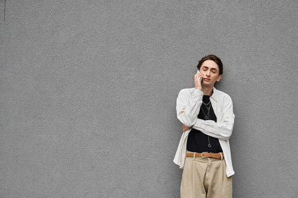 Модный молодой человек, который идентифицирует себя как педик, разговаривающий по мобильному телефону, прислонившись к серой стене.