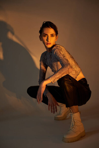A young queer person crouches in stylish attire against a white background, embodying pride.