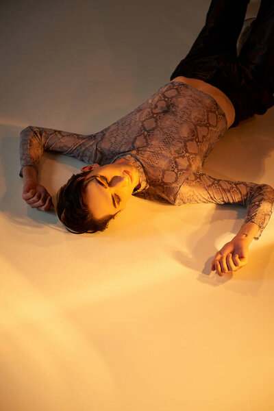 Young queer person in stylish attire lays on floor, bathed in light, exuding confidence and pride.