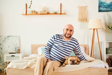 Gözlüklü bir adam, yanında Fransız Bulldog 'uyla bir yatakta huzur içinde oturur..