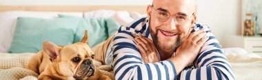 A man in glasses relaxes next to his French bulldog on a bed. clipart