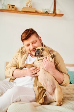 Yatakta oturan bir adam, şefkatle küçük bir köpeği tutuyor..