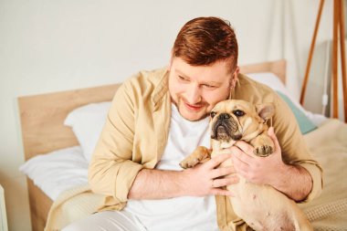 Bir yatakta oturan şık bir adam, küçük bir Fransız Bulldog 'unu sevgiyle tutuyor..