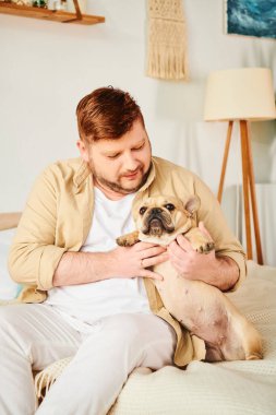 Yatakta oturan bir adam, küçük bir Fransız bulldog 'unu sevgiyle tutuyor..