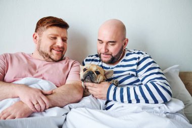 İki adam yatağın üstünde küçük bir köpek tutuyor..