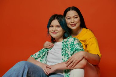 An Asian mother and her teenage daughter sit gracefully on a chair against a vibrant red background. clipart