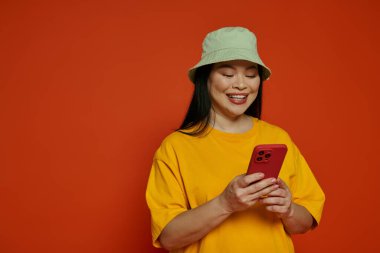 Sarı tişört giyen Asyalı kadın, turuncu arka planı olan bir stüdyoda kırmızı bir cep telefonu tutuyor..