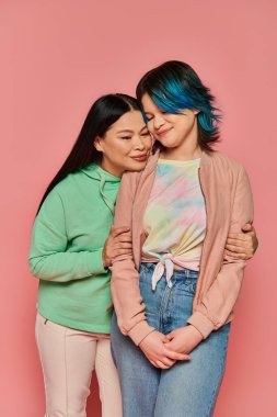 Two women, Asian mother and her teenage daughter, stand next to each other in casual wear against a vibrant pink background. clipart