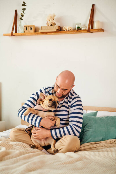 Man in glasses sitting on bed, embracing small french bulldog.