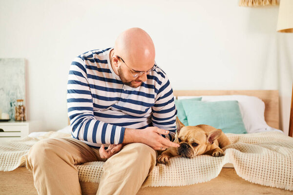 A man in glasses sits on a bed, affectionately petting a French bulldog.