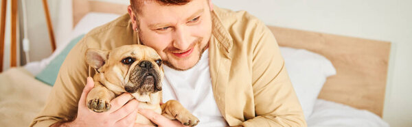 Handsome man tenderly holds a small French Bulldog in his arms at home.