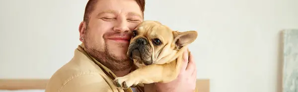 Handsome man cradling a small French bulldog in his arms at home.