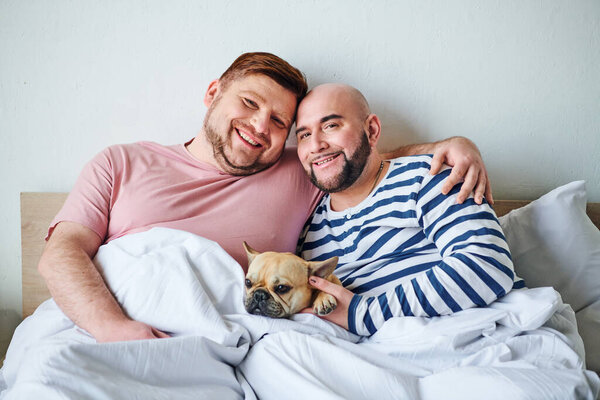 Two men cuddle in bed with their French bulldog.