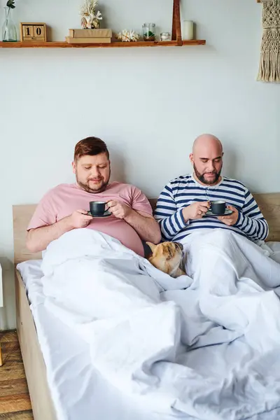 A gay couple sits together on a bed, accompanied by their French bulldog.
